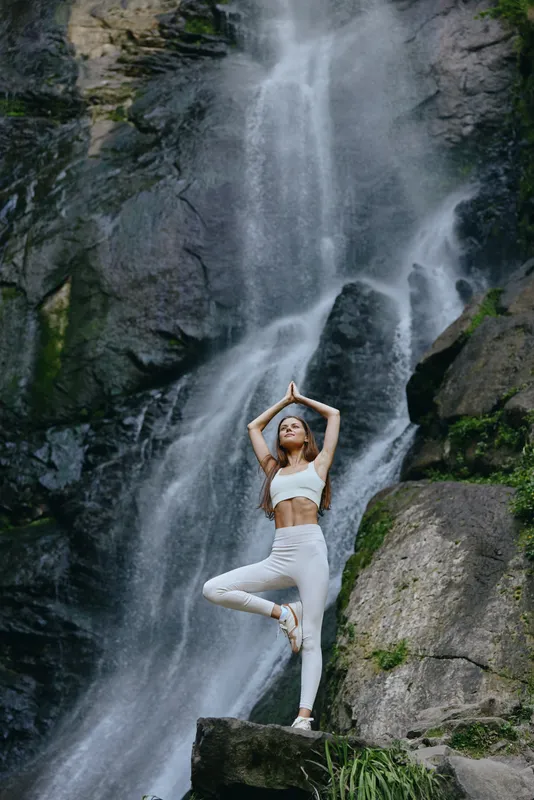 Woman practicing yoga pose near waterfall in nature, wearing white sportswear, demonstrating balance and focus with rocky natural background.