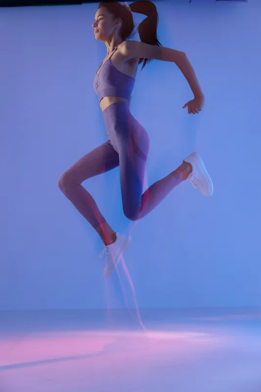 Young athletic woman sport fitness jumping mid-air in purple workout outfit with dynamic motion and energetic expression, studio lighting, vibrant blue and pink tones.