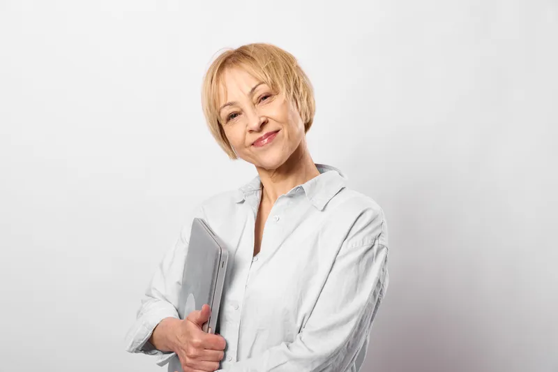 Senior woman doctor smiling in studio holds a tablet for telehealth healthcare, wearing a white coat, conveying calm guidance for online checkup in medicine