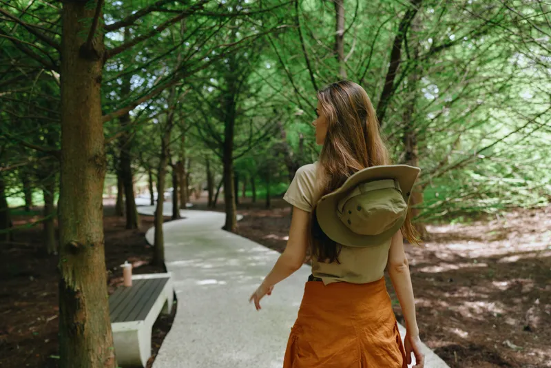 Woman walking forest path in a green park, summer casual outfit with hat carried on back, exploring shaded trees and winding trail near wooden bench for peaceful nature stroll.