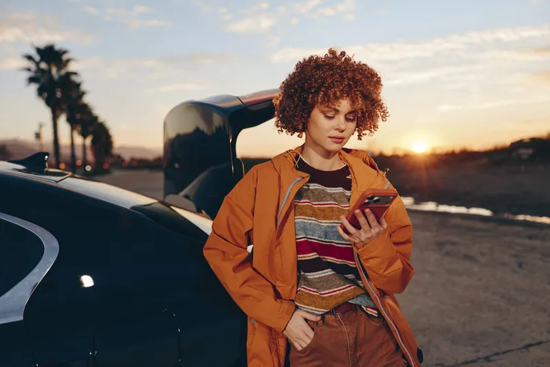 Woman lifestyle standing by car at sunset, orange jacket, curly hair, looking at smartphone, relaxed smile, outdoor street evening, travel moment, warm light, golden sky backdrop