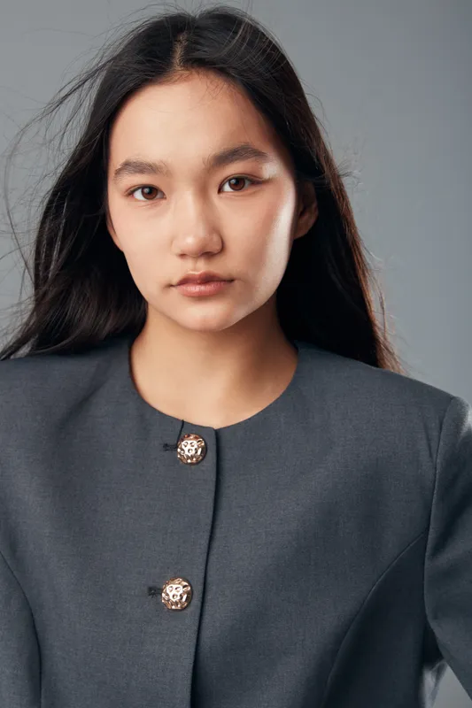 Portrait of confident young woman with straight dark hair wearing gray jacket with metal buttons looking at camera in studio. Female model with natural expression and smooth skin on neutral