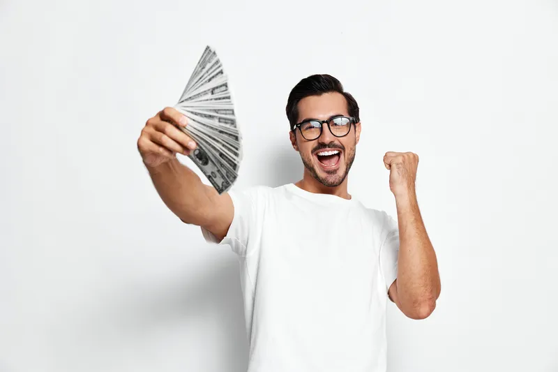 Happy smiling young man holding a fan of cash, celebrating success with excitement, wearing casual white t-shirt, isolated on plain white background.