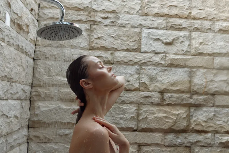 woman enjoying shower with closed eyes under rainfall in modern outdoor bathroom featuring stone wall, calming, refreshing atmosphere, natural light