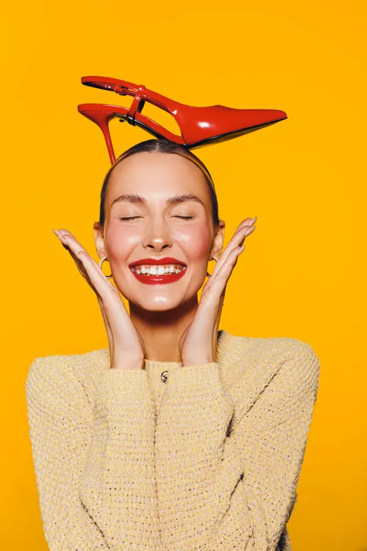 Smiling woman with closed eyes holds red high heel shoe on head against bright orange background. Concept of fashion, happiness, and playful style with vibrant colors and joyful mood.