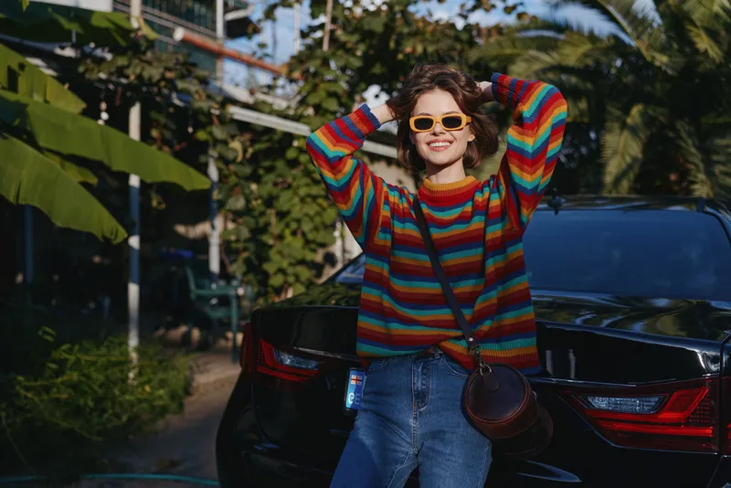 Woman smiling in a colorful striped sweater and yellow sunglasses, casual outfit and crossbody bag leaning against a black car in tropical outdoor setting, travel lifestyle vibe.