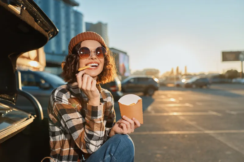 Woman sitting at car trunk eating fries in parking lot at sunset, smile with sunglasses and beanie, candid lifestyle shot with authenticity and golden hour glow, joyful urban portrait.