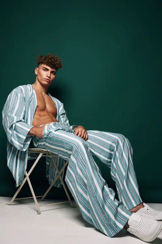 Young man sitting and posing with open shirt and striped pants in casual fashion clothes, curly hair, modern style and confident male with white slippers on chair.