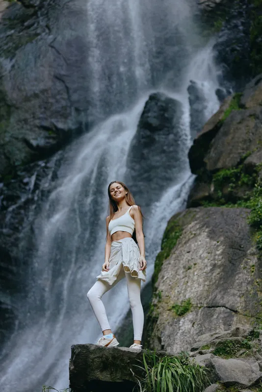 young woman standing on rock near waterfall in nature wearing white activewear, happy expression, outdoor adventure, summer season, lush greenery background