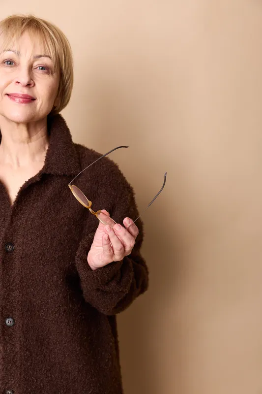 Woman portrait holding glasses, older smiling indoors, closeup near beige wall, wearing dark coat, calm expression, soft studio light, copy space