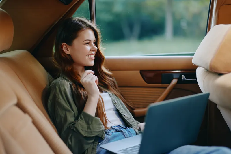 Cheerful young woman in casual outfit sitting in a car, working on a laptop with a smile, surrounded by nature in a warm, earthy color palette.