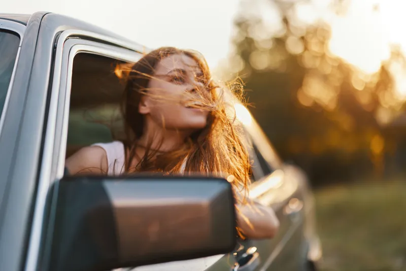 Young woman enjoying a summer road trip, with wind in her hair and a joyful expression, captured in soft golden light during sunset.