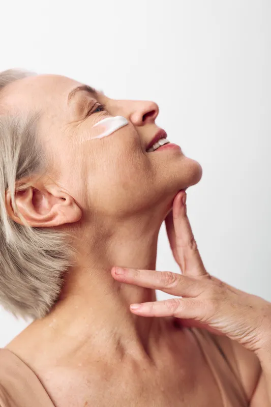 Mature woman with short grey hair applies white face cream on cheek, gently touching neck, smiling softly. Concept of skincare, aging beauty, self-care and healthy skin routine.