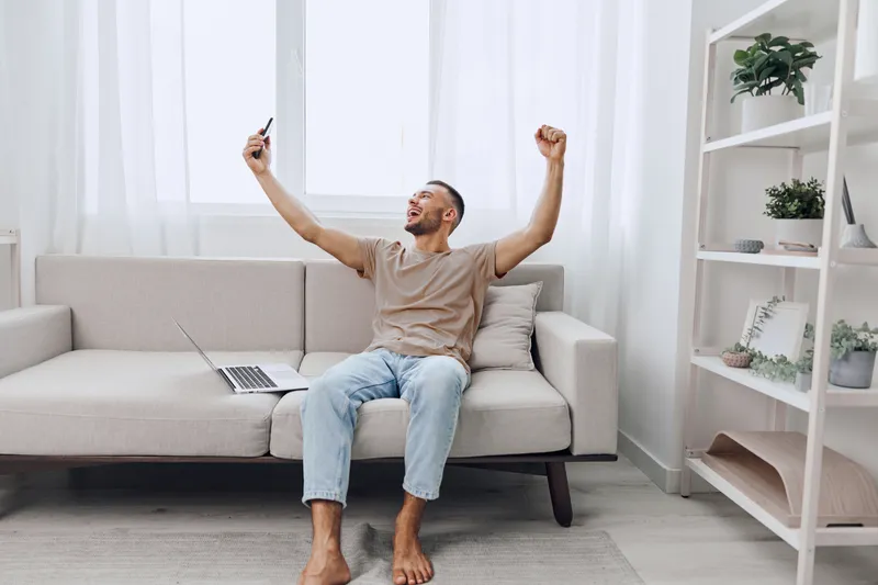 Excited man celebrating a victory at home, holding a smartphone and showing joy. Comfortable living room with natural light and a casual atmosphere.