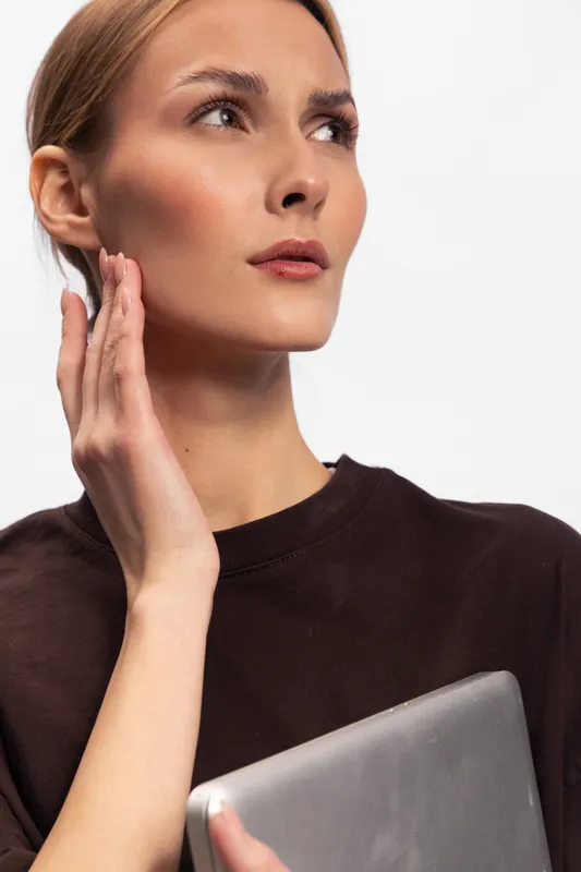 Young woman with clear skin and natural makeup looks thoughtfully aside, holding tablet in hand, wearing dark brown top against white background. Concept of technology and beauty.