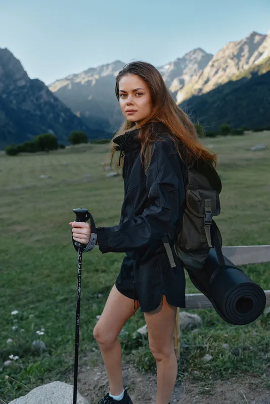 A young woman hiking in a rugged mountain landscape carries a backpack and trekking pole, ready for adventure. She smiles softly at the camera as sunlight illuminates the alpine meadow behind her.