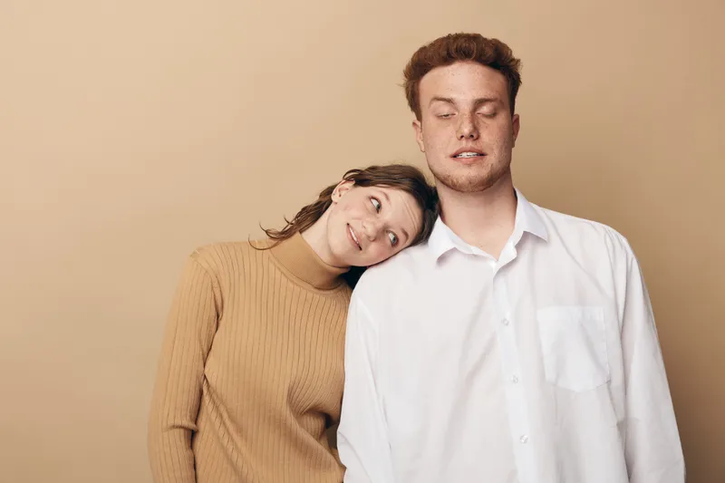 Young couple with woman resting head on man's shoulder, both with closed eyes, standing against beige background. Concept of affection, trust, and calm relationship moment.