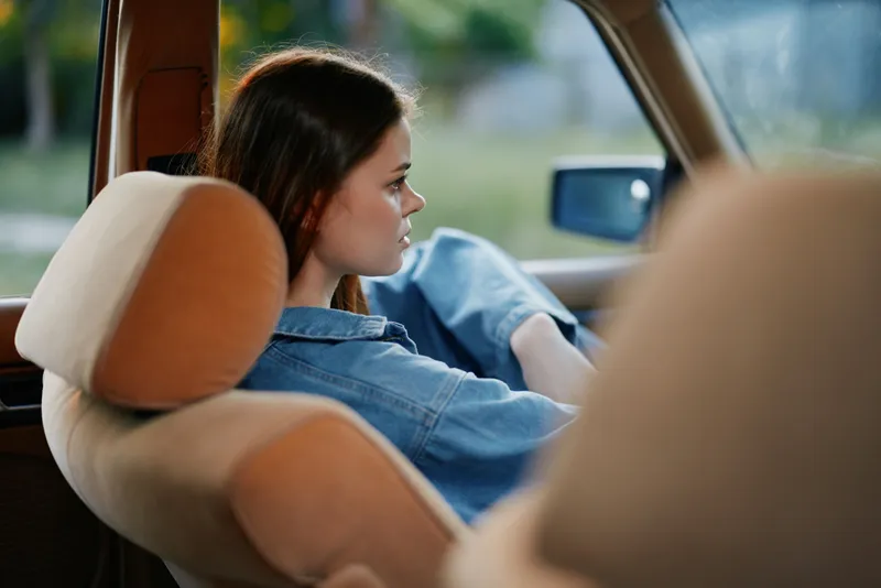 A contemplative young woman in a denim jacket relaxing in a vintage car, reflecting on life with a serene expression against a blurred natural background.