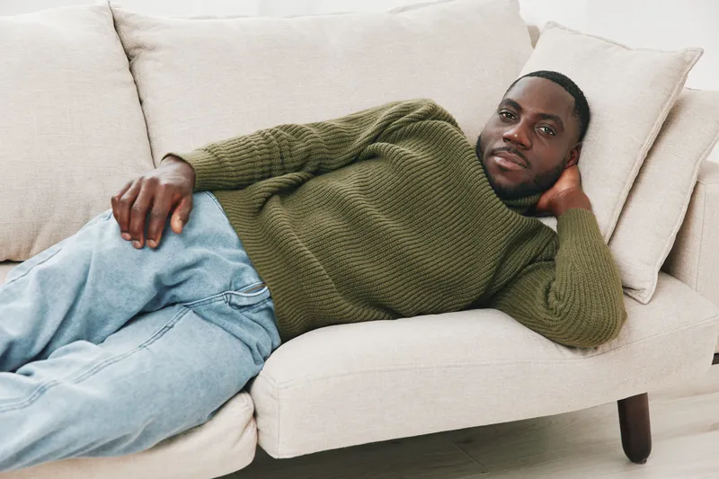 Relaxed African man lounging on a modern couch in casual green sweater and jeans, exuding calm and comfort in a contemporary living space