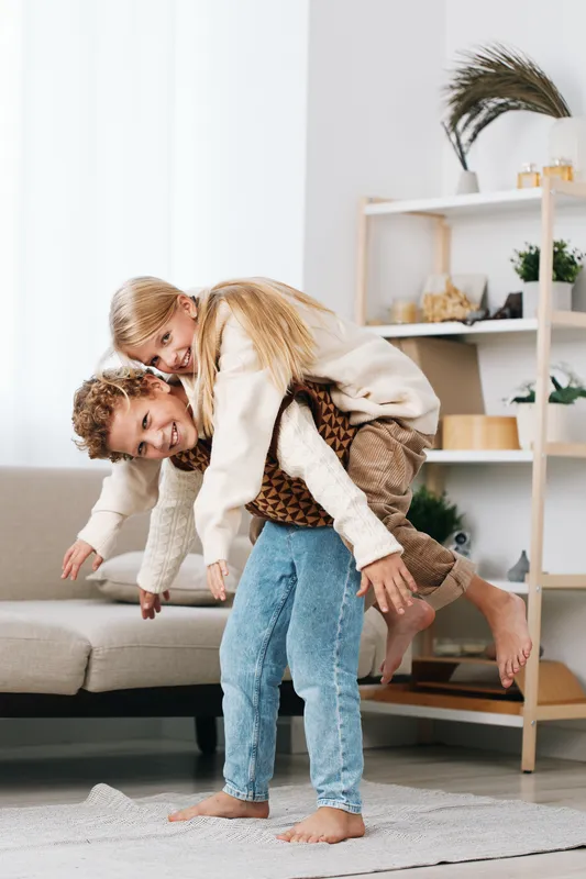 Two children playfully wrestle indoors, one climbing onto the other's back as they smile and laugh, capturing a candid moment of energetic sibling joy and carefree activity.