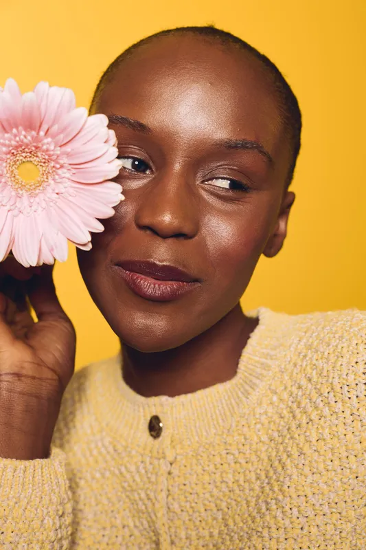 Close-up of a smiling black woman with dark skin holding a pink flower near her face against a yellow background. Concept of beauty, natural skincare, and floral wellness.