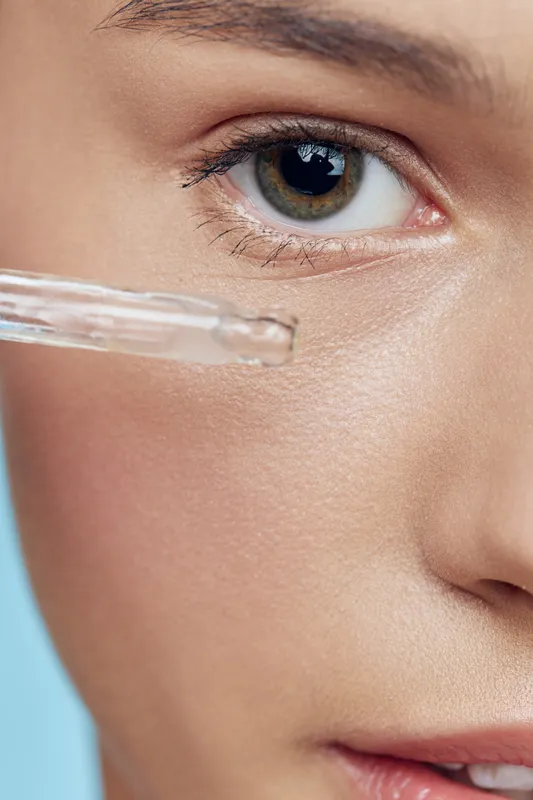 Close up of a young woman applying serum to her under eye area, showcasing healthy skincare routine with a bright background