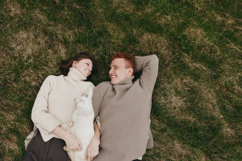 A couple wearing cozy sweaters lies on the grass, smiling at each other with a small white dog resting between them. A candid lifestyle moment that captures comfort, companionship, and simple outdoor