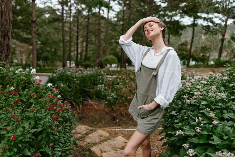 Young woman enjoying nature, wearing casual outfit with overall, smiling in a lush green garden surrounded by flowers on a sunny day, reflecting a carefree lifestyle
