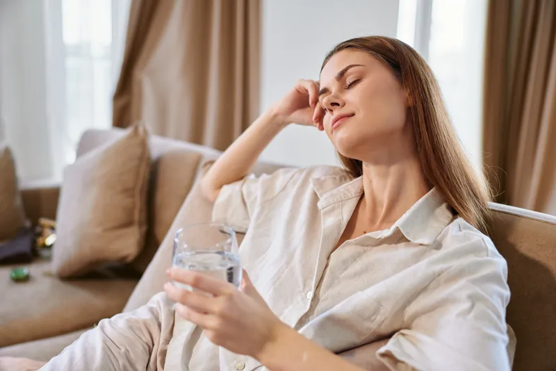 A woman enjoys relaxation on a sofa holding a glass of water at home, wellness moment in casual clothing with calm eyes closed and peaceful resting in a cozy living room.