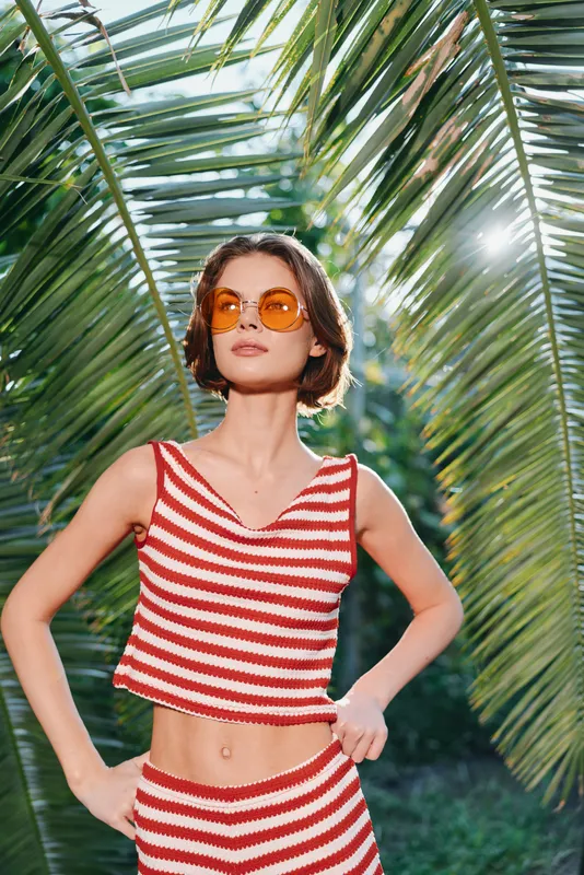 A stylish woman wearing a red and white striped crop top and matching bottoms stands under tropical palm fronds, exuding a confident summer vibe and radiant sunshine.