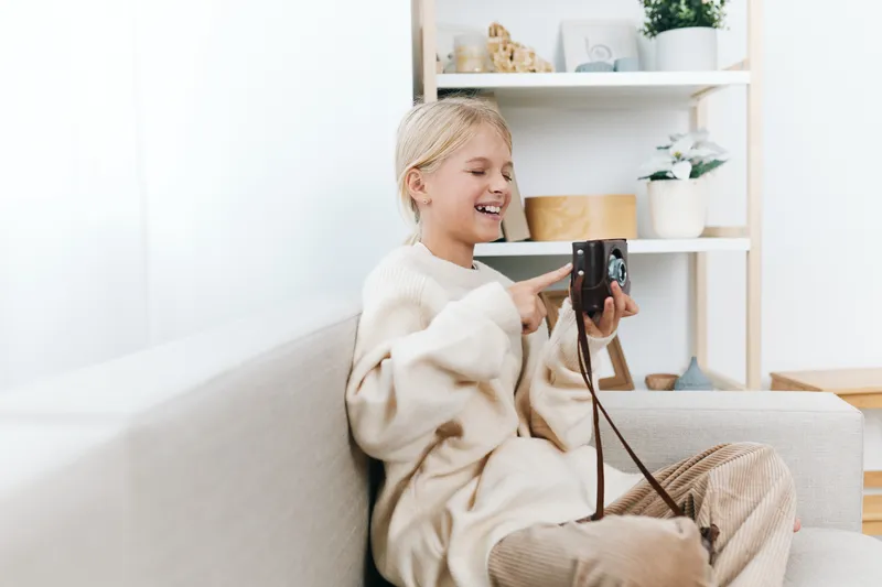 Young girl sits on a sofa, smiling as she holds a camera, capturing memories in a cozy living room with soft light, neutral tones, and relaxed vibe.