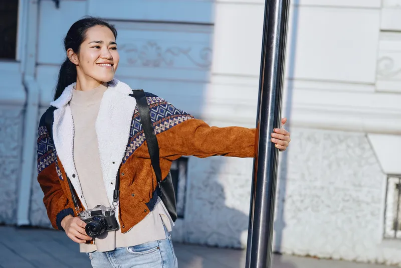 Smiling woman outdoors with a camera, wearing a brown shearling jacket and denim, backpack visible, ready for travel and photography in an urban setting, a candid moment capturing light and street
