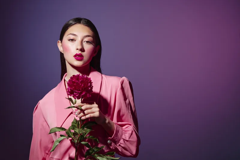 Fashion model in a pink blazer with vibrant makeup holds a red flower against purple background. Creative beauty concept with strong colors and elegant styling.