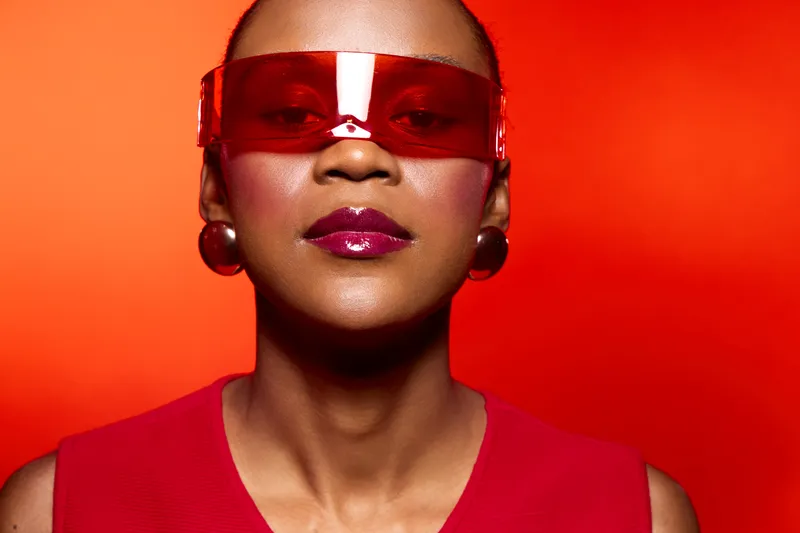 Confident African American woman with dark skin wearing red sunglasses and red outfit poses against bright red background. Concept of fashion, style, and bold modern look.