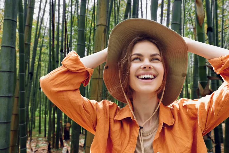Woman smile hat bamboo forest nature travel portrait: joyful female traveler in straw hat and orange shirt, laughing and looking up among tall bamboo stalks, outdoor adventure.