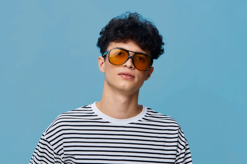 young man wearing orange sunglasses and striped shirt posing confidently against a blue background with curly hair and casual style