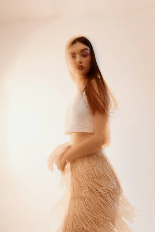 Young woman in a beige fringed skirt and white crop top poses in soft light. Fashion portrait with blurred motion effect. Concept of beauty, style, and elegance.
