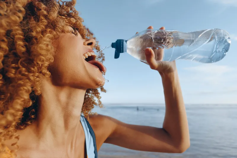 Young woman with curly hair drinks water from plastic bottle outdoors near sea under blue sky, refreshes herself after workout, summer healthy lifestyle concept.
