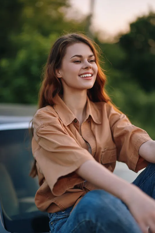 Smiling young woman in casual brown shirt and jeans enjoying a sunny day outdoors, surrounded by lush greenery.