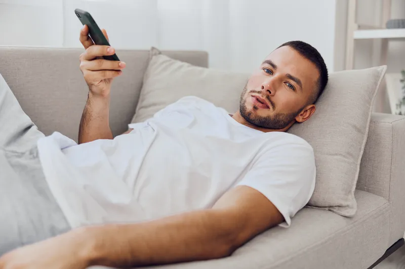 Relaxed man lying on a sofa, using a smartphone while enjoying his free time at home. Modern and cozy interior with a calming atmosphere makes it inviting.