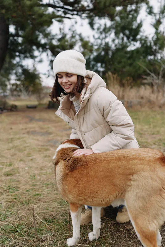 Woman in a warm winter coat and beanie outdoors, gently touching a friendly dog in a grassy field on a chilly day, capturing a candid moment of companionship and nature.
