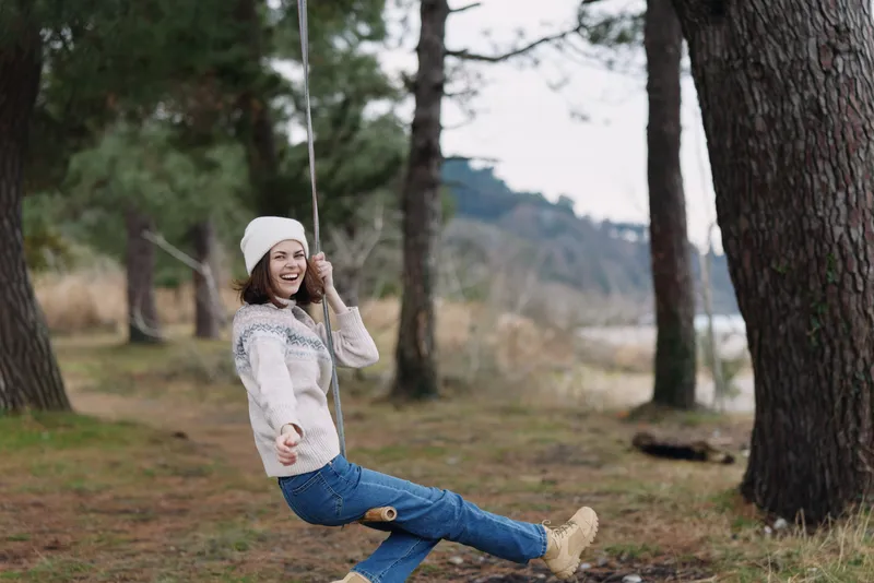 Woman on a swing in a forest, smiling and enjoying a breezy outdoor moment, casual sweater and jeans, peaceful nature scene for lifestyle and leisure stock