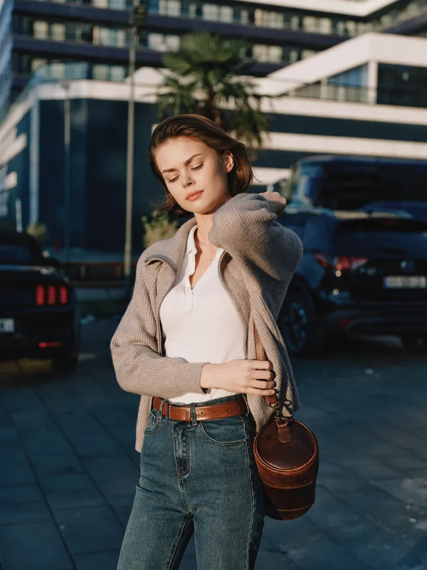 A thoughtful young woman stands outdoors near a modern building, with eyes closed and hand brushing her hair, conveying reflection and calm amid a busy urban setting.