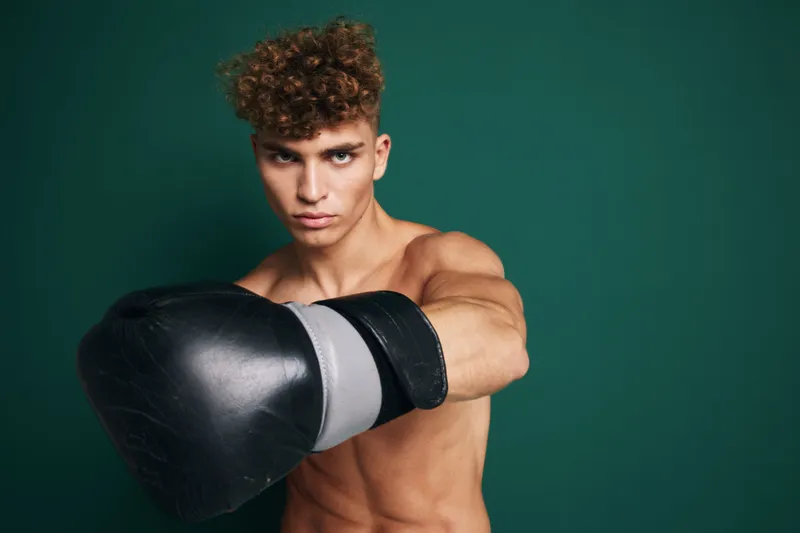 Young male boxer with curly hair wearing black boxing gloves in fighting stance against green background. Muscular athlete training for boxing or martial arts workout.