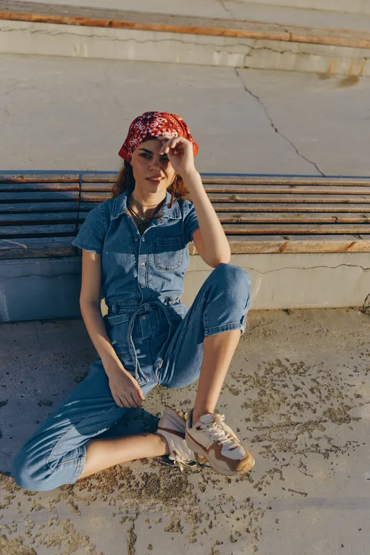 lifestyle woman in modern boho-western denim street style with red bandana sits outdoors on urban pavement, casual relaxed pose, natural light, film color tones