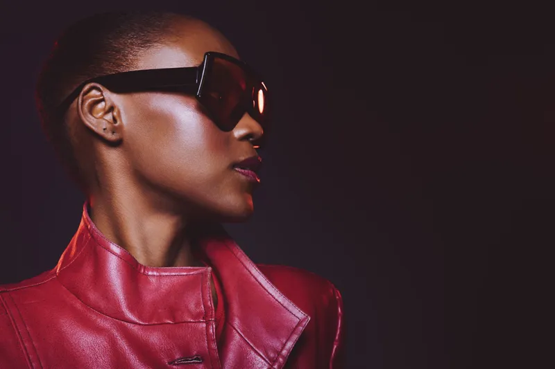 Stylish dark skinned African woman wearing large sunglasses and a red leather jacket poses in profile against dark background. Concept of fashion, elegance, and confidence.