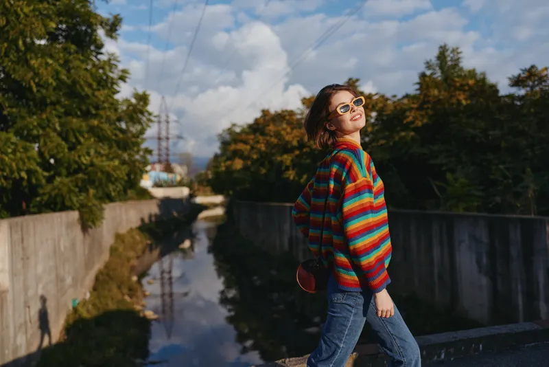 Woman in a colorful striped sweater and sunglasses walking on an urban bridge, smiling and leaning back in casual jeans with a red crossbody bag for travel lifestyle.
