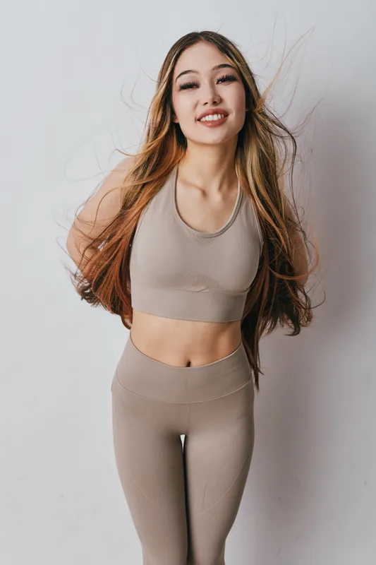 Young woman in beige sportswear smiling with long hair flowing, posing for fitness portrait on white background. Healthy lifestyle, active, confident, and happy female athlete in studio.