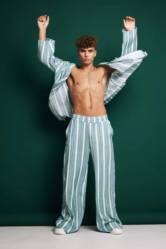 Young man with curly hair wearing striped pajamas posing with raised arms against dark green background. Fashion, casual, relaxed, confident, studio portrait with modern style.