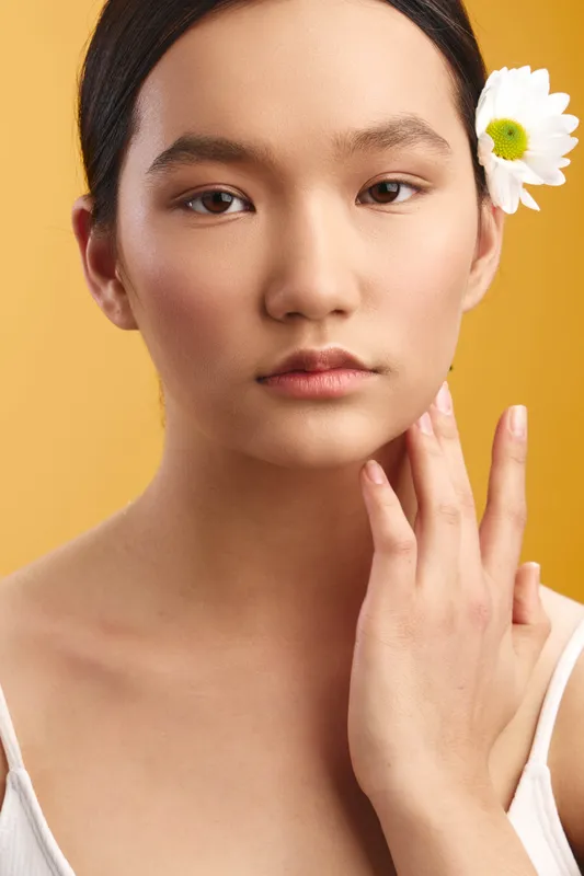 asian woman with a flower tucked behind her ear smiles softly, a calm presence in a warm yellow studio, highlighting natural beauty and confident poise.